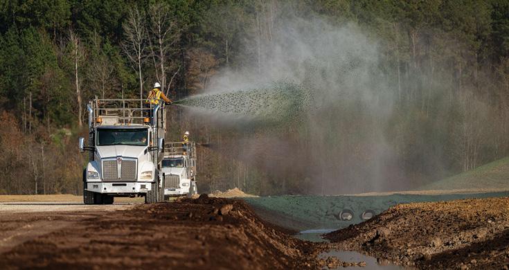 A worker on a truck sprays a hydromulching grass seed mixture on a roadside slope with another truck behind.