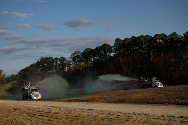 Two trucks spray green hydromulching material on dirt beside a road, with trees and blue sky in the background.
