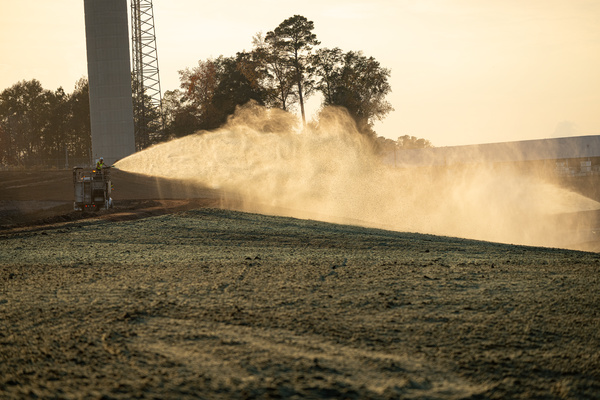 A truck sprays hydroseeding slurry over a grassy field near trees and a tall structure at sunrise or sunset.