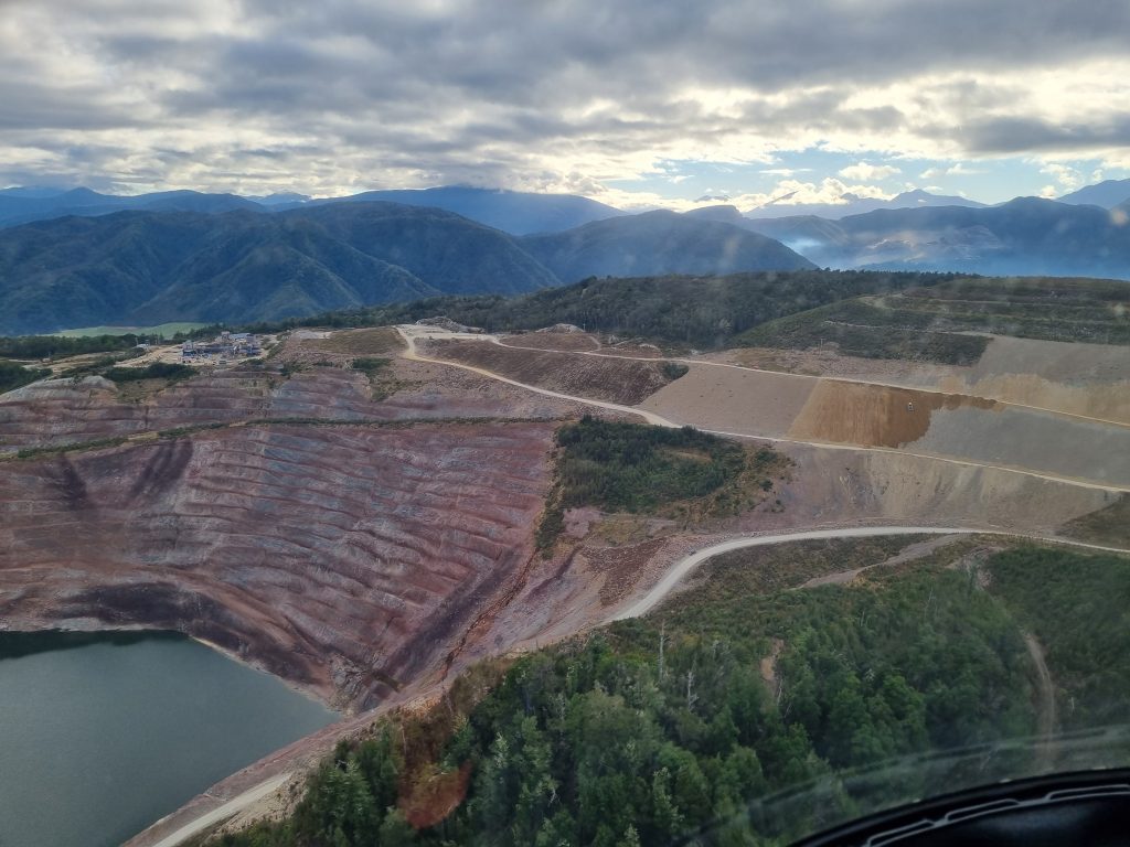 Aerial view of a large open-pit mine with terraced walls and a reservoir, set against a mountainous landscape.