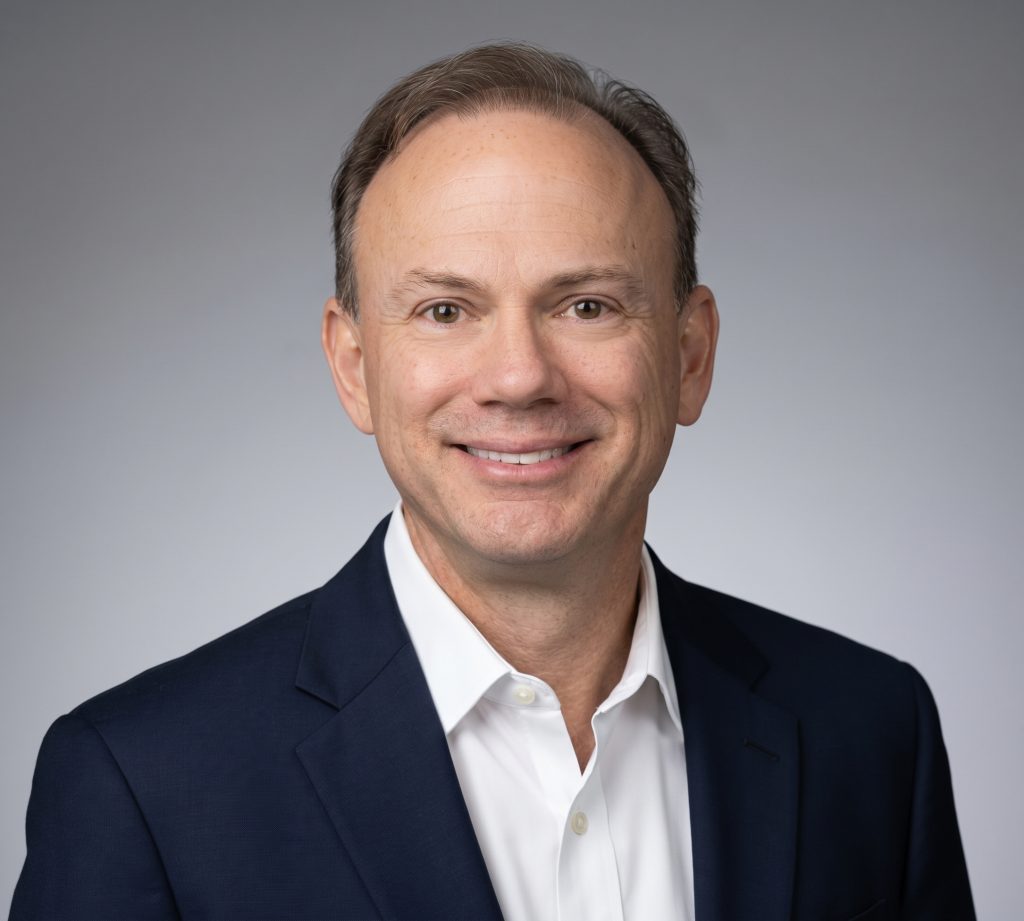 Smiling man in a dark suit jacket and white shirt against a plain light gray background.