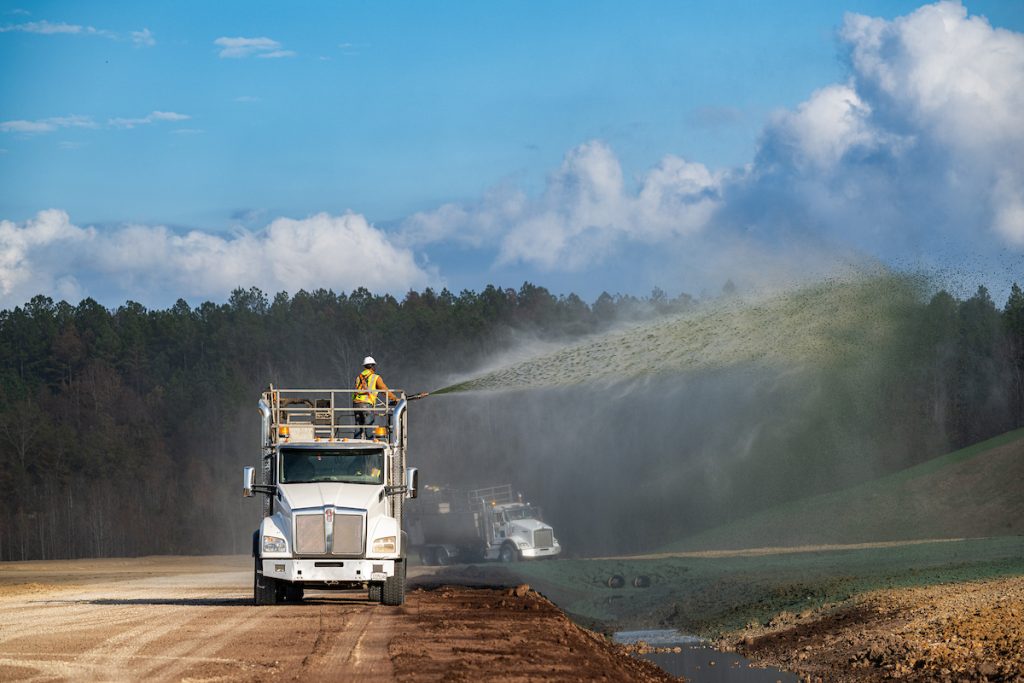 A worker on a truck sprays a green mixture onto a roadside embankment.